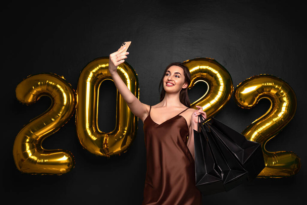  Brunette woman in elegant dress hold shopping bags doing selfie shots on smartphone on black background, numbers air balloons studio. New Year shopping ,Black Friday, sales. 2022 - 写真・画像