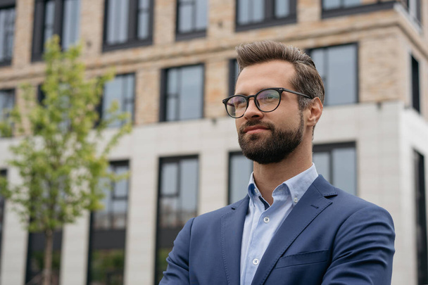 Portrait of confident businessman wearing suit and stylish eyeglasses standing outdoors. Young handsome manager planning project, looking away at copy space. Successful business, career - Foto, Imagen