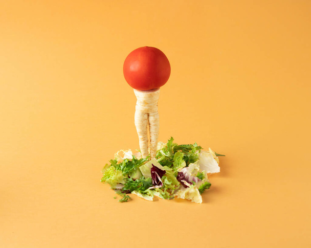 Parsley root like a woman's legs and red tomato standing on mixed salad on yellow background. Minimal food arrangement. - Foto, imagen