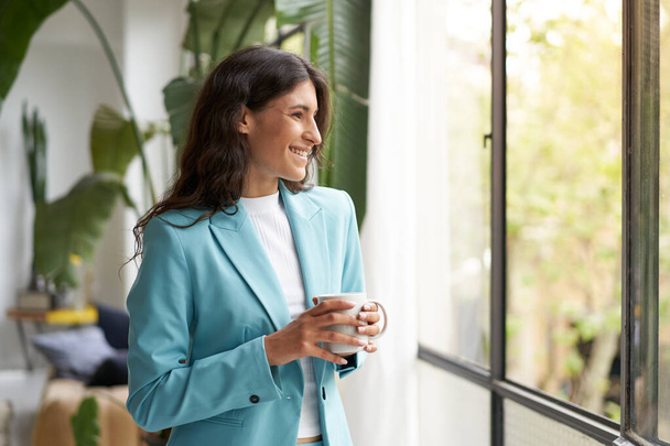 Jeune femme d'affaires souriante satisfaite d'un travail bien fait relaxant avec son café ou son thé du matin, regardant par la fenêtre. Belle latina femme célébrant les succès dans le bureau. - Photo, image