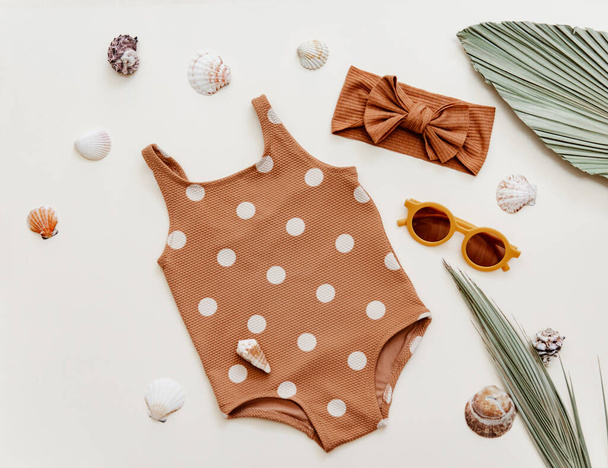 Little girl's cute swimsuit with sunglasses and seashells on beige background. Retro fashion baby, summer holidays concept - Photo, Image