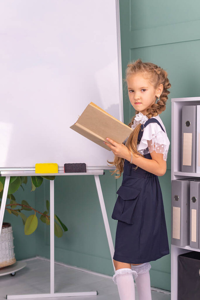 Back to school and happy time! A cute hardworking child is sitting at a table indoors. girl studying in the classroom on the background of the blackboard. The girl is reading a book. - Foto, Imagen