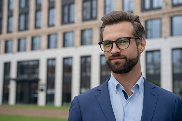 Portrait of attractive pensive banker looking at camera, copy space. Young confident businessman wearing suit, stylish eyeglasses standing on the street. Successful business and career concept - Fotó, kép