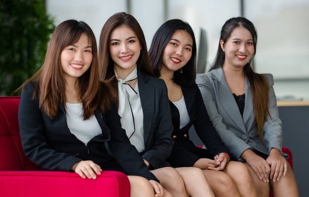 Grupa czterech atrakcyjnych azjatyckich koleżanek z biura w formalnych garniturach, siedzących na czerwonej kanapie w biurze i uśmiechających się do kamery. Pojęcie nowoczesnej pracy biurowej. - Zdjęcie, obraz