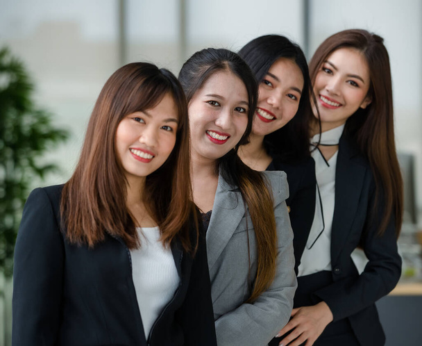 Ομάδα από τέσσερις ελκυστικές Ασιάτισσες συναδέλφους γραφείου με επίσημα κοστούμια που στέκονται στο γραφείο χαμογελώντας στην κάμερα. Έννοια για σύγχρονο γραφείο εργασίας. - Φωτογραφία, εικόνα