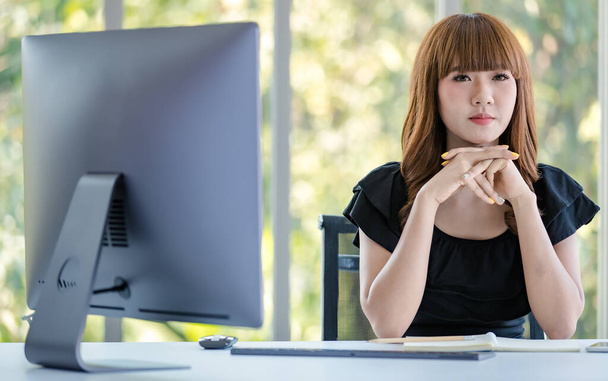 Azjatycka bizneswoman wspierająca podbródek obiema rękami i uśmiechnięta do kamery, komputer blisko niej, koncepcja pracy w biurze. - Zdjęcie, obraz
