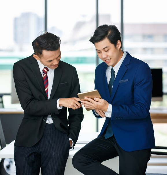 Empresários asiáticos positivos em ternos em pé no local de trabalho e tablet de navegação juntos durante o trabalho no projeto de negócios - Foto, Imagem