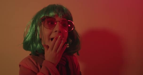 Mała dziewczynka dmuchanie pocałunek noszenie kolorowe peruki i okulary przeciwsłoneczne w kształcie serca pozowane do fotografowania na tle światła disco - Materiał filmowy, wideo