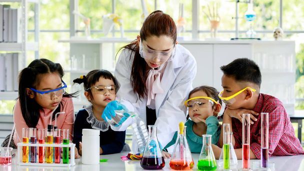 白衣ゴム手袋と安全眼鏡のアジアの女性専門家の科学教師は、試験管顕微鏡で実験室で小学生や女子学生に科学実験を教えています. - 写真・画像