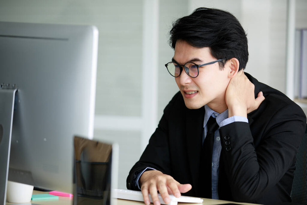 Ázsiai fiatal fekete rövid haj férfi alkalmazott munkavállaló visel formális öltöny nyös szürke ing és szemüvegek irodai szindróma fáj a váll és a nyak, amikor dolgozik elülső számítógép monitor asztalnál. - Fotó, kép