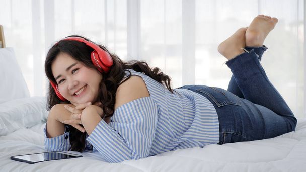 Asiatische junge glückliche ziemlich freundliche dicke übergroße Frau legte sich lächelnd entspannt auf ein weißes Bettlaken und hörte Musik aus großen roten Kopfhörern.. - Foto, Bild