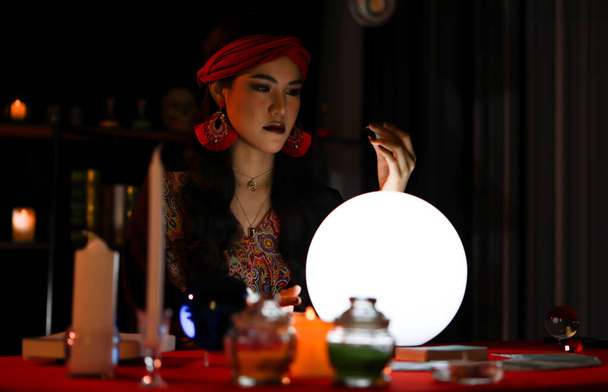 Eine schöne Frau mit langen Haaren und rotem Turban agiert als Wahrsagerin in einem dunklen Raum - Foto, Bild