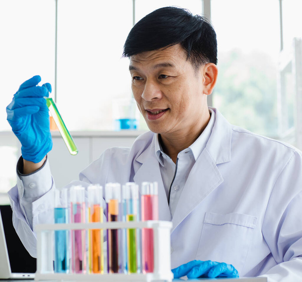 2 asian men are scientist holding and look at test tube with chemical liquid. Focus to young scientist. Microscope, laptop and more test tubes with liquid put on table in laboratory room in the morning. - Photo, Image