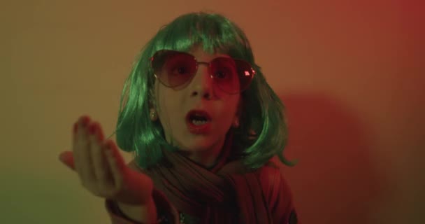Klein meisje blazen kus het dragen van een kleurrijke pruik en hartvormige zonnebril poseerde voor een foto schieten op de disco lichte achtergrond - Video