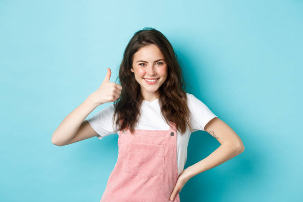 Portret van zelfverzekerde en positieve jonge vrouw duim omhoog, ja zeggen, toestemming geven, goedkeuren en eens iets goeds, lof mooie keuze, staande tegen een blauwe achtergrond - Foto, afbeelding