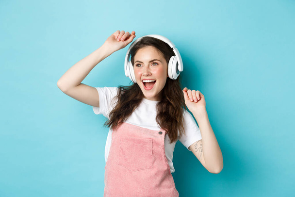 Imagem de menina bonita alegre dançando em fones de ouvido, ouvindo música no aplicativo e pulando animado, de pé sobre fundo azul - Foto, Imagem