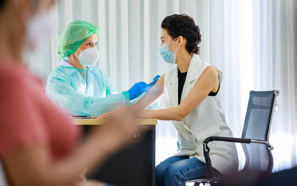 白人女性プロの医師は、デスクで若い女性患者の肩に注射針注射コロナウイルスワクチンを使用して完全なPPE保護制服顔マスクゴム手袋ラボコートと帽子を身に着けています. - 写真・画像