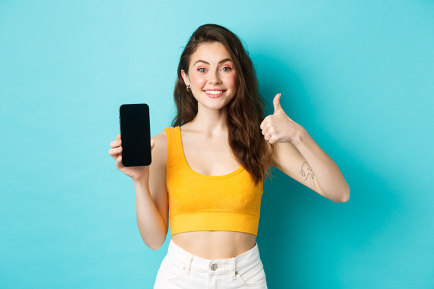 Ritratto di elegante donna moderna sorridente soddisfatto, raccomandare app, mostrando banner logo sullo schermo dello smartphone vuoto, facendo pollice in su in approvazione, lode applicazione, sfondo blu - Foto, immagini