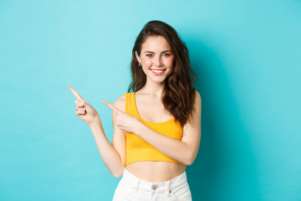 Portré elegáns lány színes nyári ruhában, ujjal balra mutató promóciós ajánlat, mutatja a logó, mosolygós boldog a kamera, áll a kék háttér felett - Fotó, kép