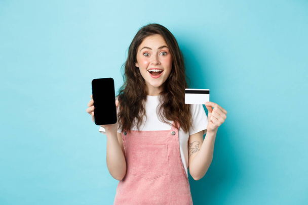 Смотри сюда. Возбужденная молодая милая девушка показывает пустой мобильный экран и пластиковые кредитные карты, смотреть поражены камерой, стоя на синем фоне - Фото, изображение