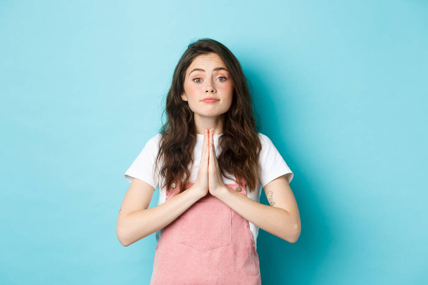 Please help me. Cute brunette woman asking for favour, holding hands in pray, begging you to buy something, standing against blue background - Fotó, kép