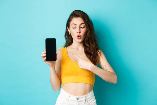 Young excited woman in summer cropped top, pointing and looking at empty smartphone screen, showing app or online shop, standing over blue background - Фото, изображение