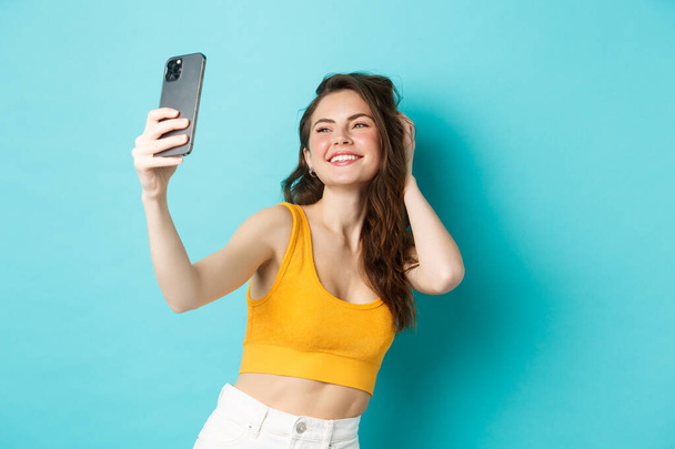 Mooi modern meisje nemen selfies op vakantie, poseren in de zomer kleren en kijken naar smartphone camera, staande tegen een blauwe achtergrond - Foto, afbeelding