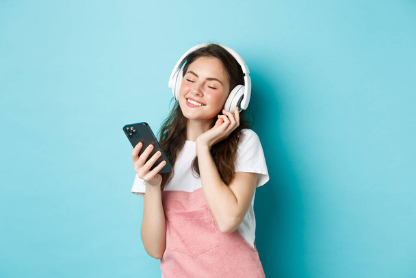 Piękna kobieta ciesząca się piosenką w słuchawkach, zamkniętymi oczami i uśmiechem słuchając muzyki w słuchawkach, trzymając smartfona w ręku, stojąc nad niebieskim tłem - Zdjęcie, obraz