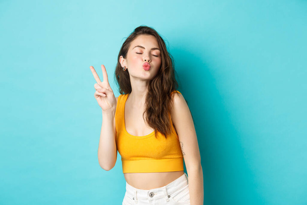 Glamour dívka v letním oblečení zavřít oči a dělat líbání tvář s V-znamení, těší dovolenou, baví, stojí uvolněně na modrém pozadí - Fotografie, Obrázek