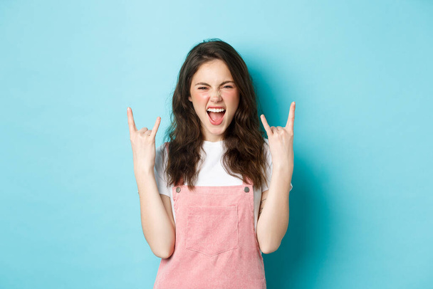 Portrait of excited woman enjoying concert or awesome event, showing rock n roll horns gesture and shouting with joy, having fun, standing over blue background - Photo, Image