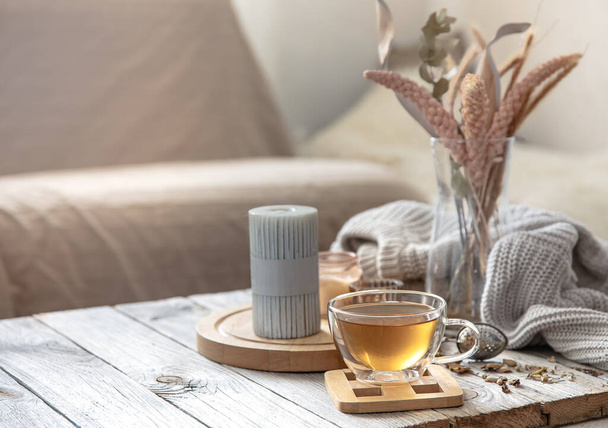 Acogedora composición casera de otoño con una taza de té, velas y un elemento de punto sobre un fondo borroso del interior de la habitación. - Foto, imagen