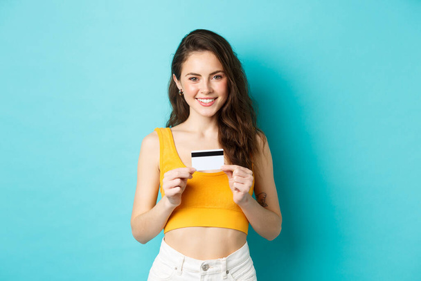 Ψώνια. Χαμογελώντας όμορφη γυναίκα σε καλοκαιρινά ρούχα, δείχνει πλαστική πιστωτική κάρτα, αναζητούν αυτοπεποίθηση, στέκεται πάνω από το μπλε φόντο - Φωτογραφία, εικόνα