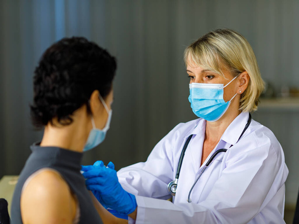 Beautiful short black hair female patient wears face mask sit look at camera while Caucasian doctor in white lab coat with stethoscope injecting vaccine in to her shoulder in blurred background. - Фото, зображення