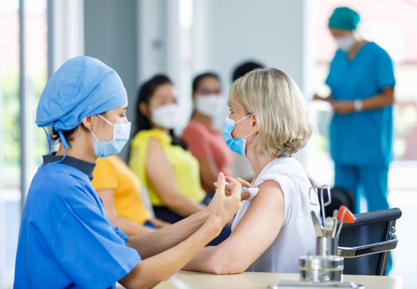 Női hivatásos orvos visel arc maszk és kék kórházi egyenruha befecskendezés coronovírus vakcina fecskendő tű a kaukázusi idős nő beteg váll klinika kórterem munkaasztal. - Fotó, kép