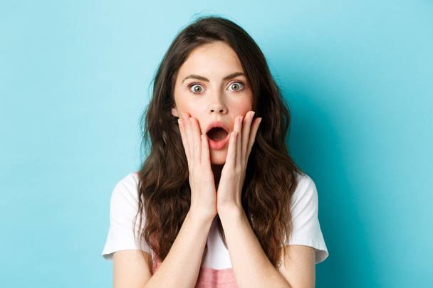 Oh my gosh. Close up portrait of shocked girl face, touching head and staring at camera startled, hear bad news, standing over blue background - Photo, Image
