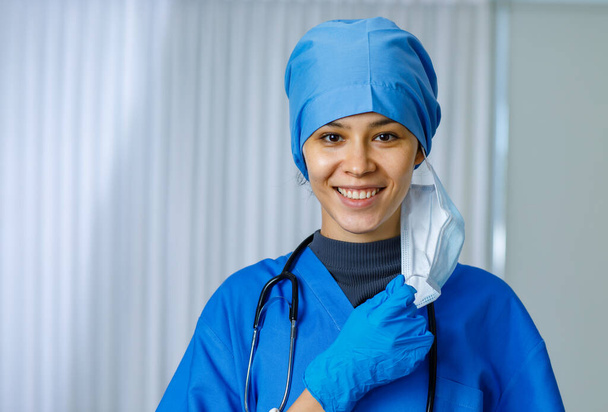 Портрет крупним планом зйомка щасливої красивої жінки-лікар в блакитному лікарняному костюмі зі стетоскопом, який посміхається, коли знімає використану хірургічну маску для обличчя, дивлячись на камеру, коли закінчується пандемія коронавірусу
. - Фото, зображення