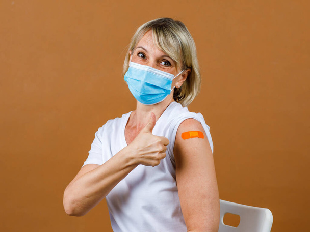 Beyaz tenli, sarışın, yüz maskesi takan bir hastanın portresi. Coronavirus aşısı olduktan sonra omzundaki turuncu bandaj başparmağını gösteriyor.. - Fotoğraf, Görsel