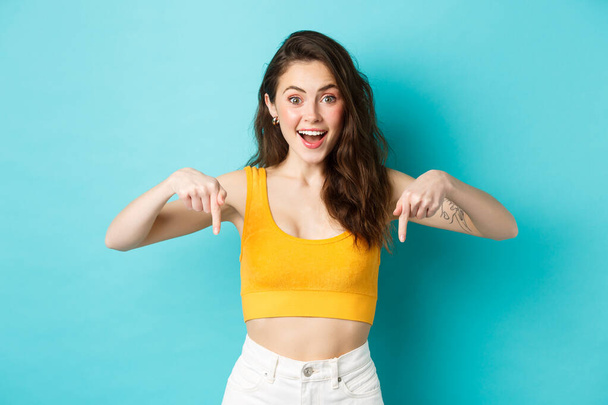 Young happy woman showing awesome news, pointing fingers down, demonstrate advertisement on bottom smiling excited, standing against blue background - Zdjęcie, obraz