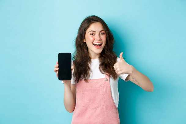 Yes this is good. Smiling cute girl showing thumb up and empty smartphone screen, recommending application or online store, standing over blue background - Fotoğraf, Görsel