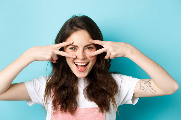 Close up of attractive excited woman showing v-signs, disco gesture near eyes, smiling pleased, standing upbeat against blue background - Photo, Image