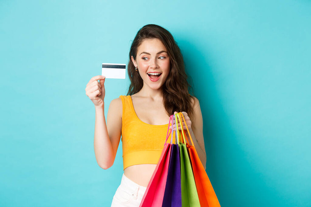 Image d'une femme heureuse accro au shopping montrant sa carte de crédit en plastique, tenant des sacs à provisions, portant des vêtements d'été, debout sur fond bleu - Photo, image