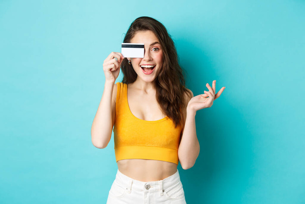 Excited beautiful woman in summer cropped top, look amazed at camera, shopping with credit card, standing over blue background - Photo, Image
