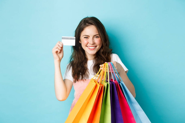 Joven mujer satisfecha sonriendo, mostrando tarjeta de crédito de plástico y la celebración de bolsas de compras, compra con el pago sin contacto, de pie sobre fondo azul - Foto, Imagen