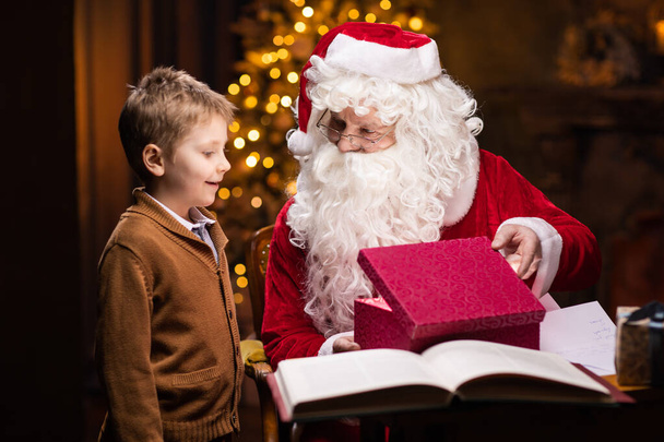 Santa Claus and little boy. Cheerful Santa is working while sitting at the table. Fireplace and Christmas Tree in the background. Christmas concept. - Foto, Imagem
