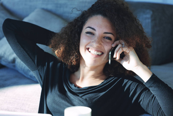 Iloinen nuori nainen nauraa ja hymyilee, kun hän keskustelee ystävänsä kanssa matkapuhelimellaan rentoutuessaan lattialla kotona kesällä auringonpaisteessa - Valokuva, kuva