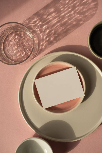 Leere Papierbögen mit Kopierattrappe auf staubig rosa Hintergrund. Ästhetisch unkonventionelle Geschäftsmarken-Vorlage. Keramische Teller und Glas mit glitzernden Sonnenschatten - Foto, Bild