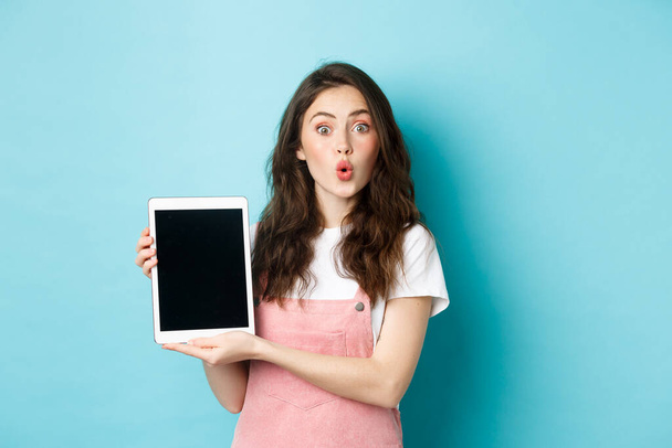 Verrast aantrekkelijke vrouw tonen digitale tablet scherm en kijken verbaasd op camera, controleren promo aanbod, staande geamuseerd tegen een blauwe achtergrond - Foto, afbeelding