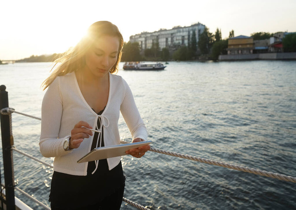 serious business woman in summer outfit standing near coast, using tablet with pen,  looking at the screen. Outdoor shot, sunset. Work on vacation or freelance concept - Valokuva, kuva