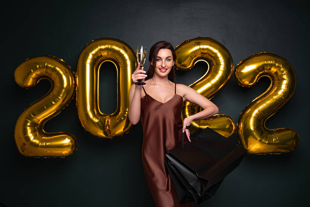 Beautiful brunette model stands on a dark background with air balloons letters 2022 and holds glass of champagne, shopping bags, gifts. confetti fly around, merry Christmas and happy new year - Foto, afbeelding