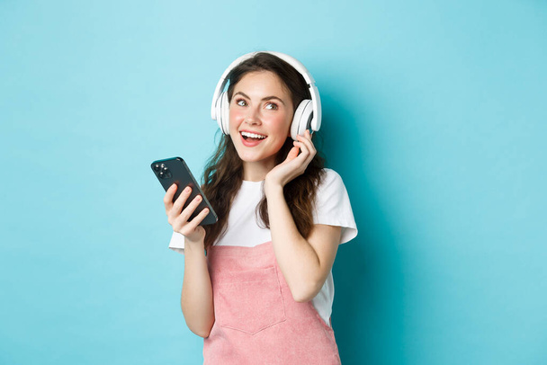 Bild einer jungen Frau, die Kopfhörer aufsetzt und Musik auf dem Smartphone auswählt, das Telefon hält, Song hört, vor blauem Hintergrund steht - Foto, Bild